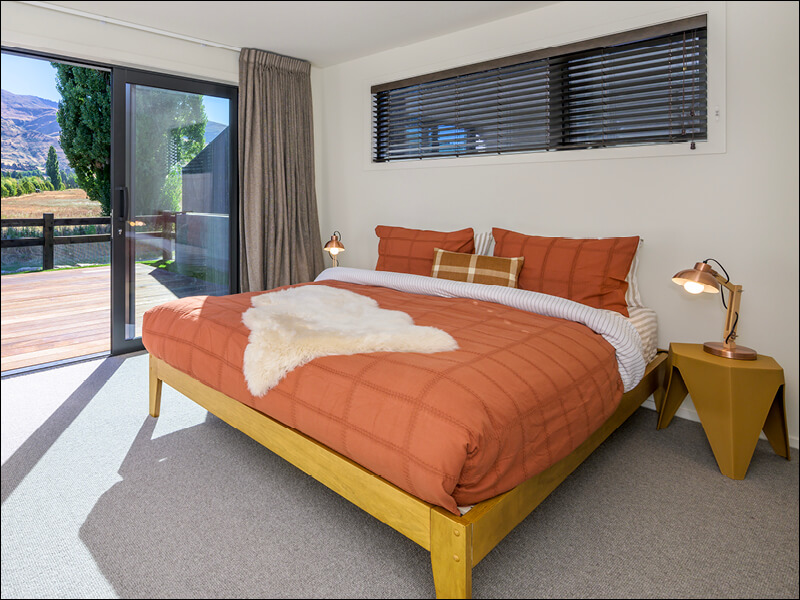 Modern bedroom in Wanaka home with orange bedding, wooden bed frame, bedside tables, and sliding glass door opening to a private deck