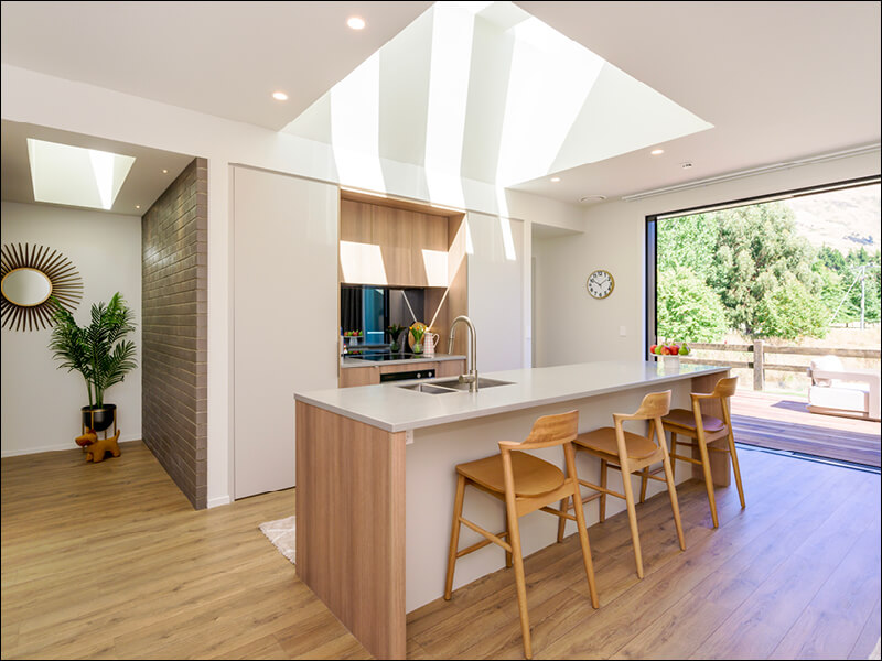 Bright open-plan kitchen and dining area in a Wanaka home featuring skylights, wooden flooring, and large sliding doors opening to a deck.