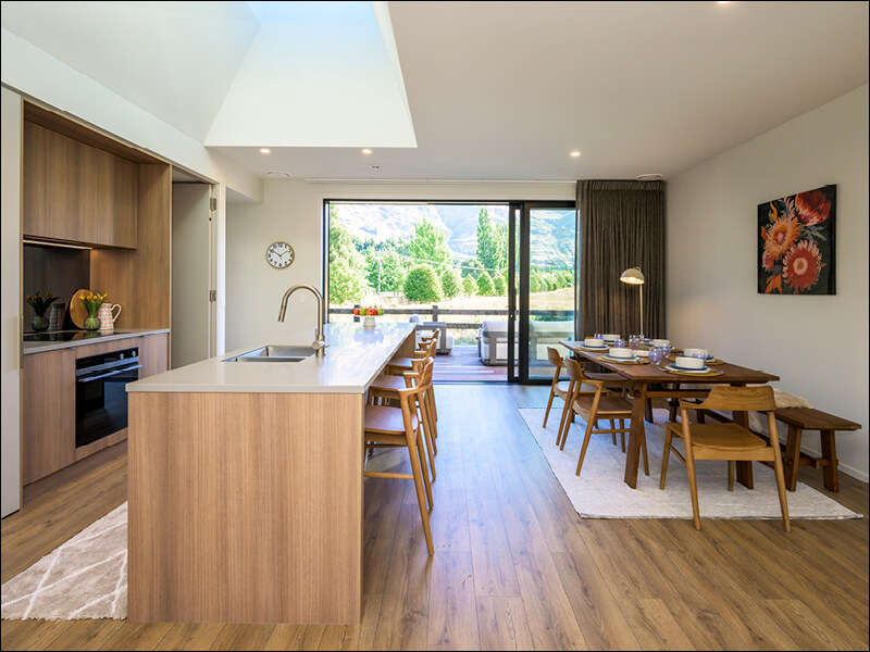 Open-plan kitchen and dining area in a modern Wanaka home, featuring wood cabinetry, a large island, and sliding glass doors with mountain views