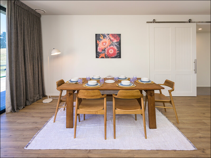 Contemporary dining room with wooden table set for six, mid-century chairs, white rug, and floral artwork in a Wanaka house