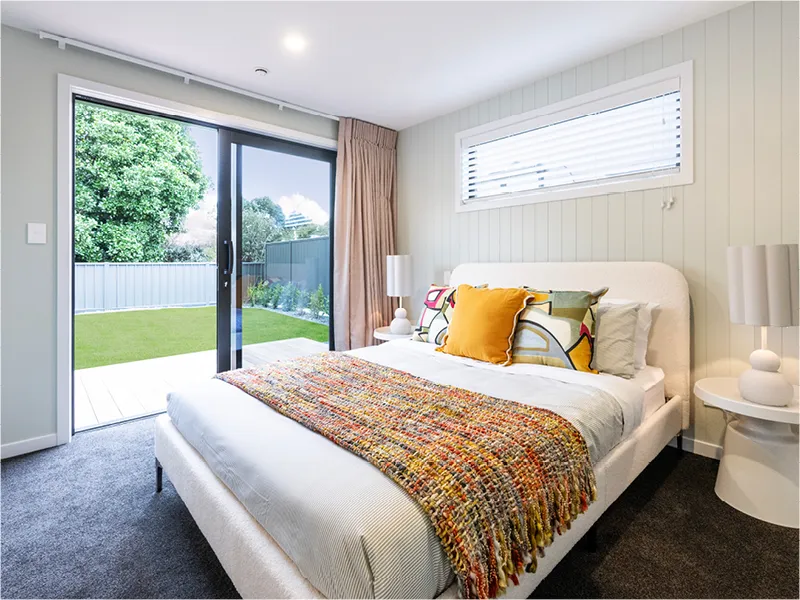 Master bedroom in the Napier lottery home featuring a light upholstered bed, colourful throw and cushions, and sliding glass doors opening to a landscaped backyard.