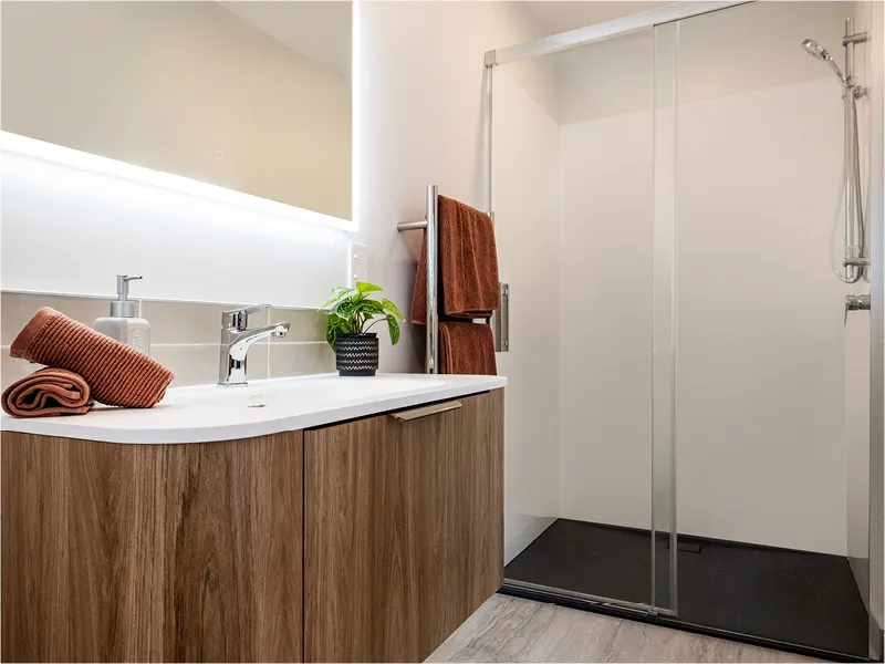 Modern bathroom vanity with timber cabinet, LED backlit mirror and towel rail, viewed from bedroom doorway.