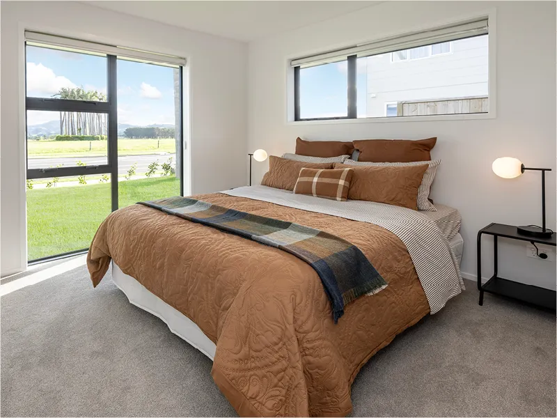 Bright modern bedroom with large window, brown bedding, bedside tables and lamps, and soft neutral carpet