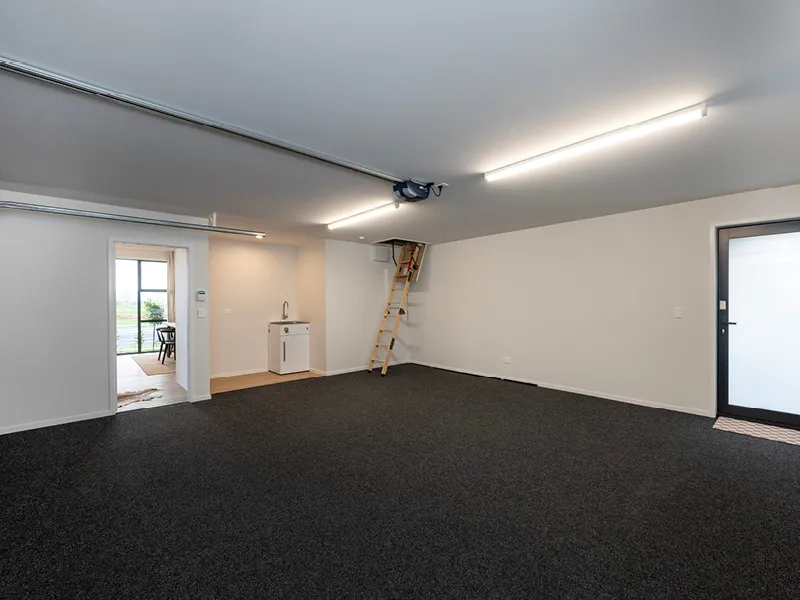 Spacious double garage interior with dark carpet flooring, internal access door, laundry sink and attic ladder storage.