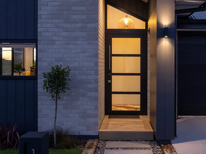 Front door entry close-up with modern glass-panel door, exterior lighting and brick wall at dusk