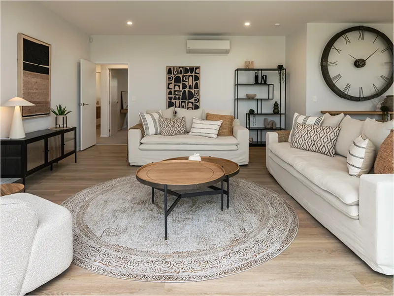 Open-plan living room with two light sofas, round timber coffee table, large wall clock and neutral decor