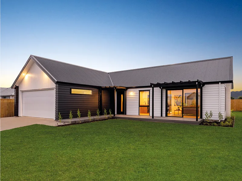 Modern single-storey Whitianga home with gable roof, double garage and large green lawn at sunset, exterior lights glowing through floor-to-ceiling windows.