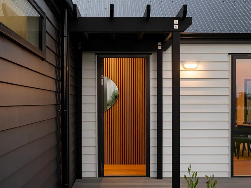 Modern Whitianga home entry with black pergola, white weatherboard cladding and feature front door with vertical timber slats and circular cut-out detail at dusk.
