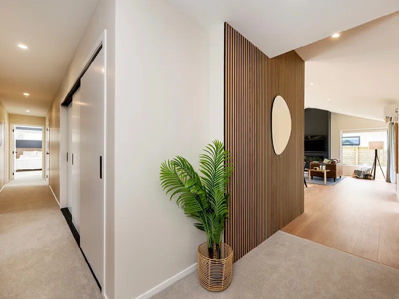 Modern Whitianga home interior hallway with sliding storage doors, feature timber slat partition with oval mirror, potted plant and open-plan living area with timber flooring in the background.