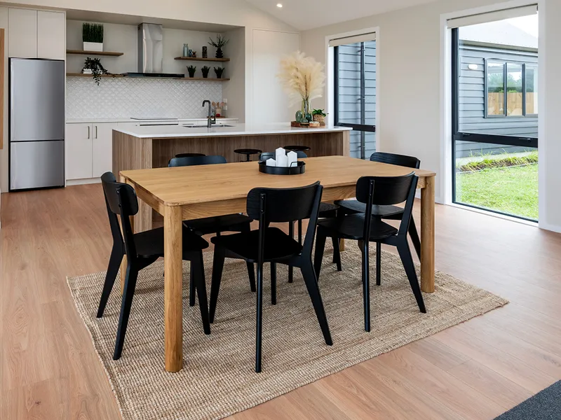 Modern Whitianga open-plan dining area with wooden table and black chairs on a textured rug, adjacent to contemporary white kitchen with island bench and large windows to the backyard.