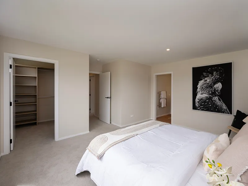 Modern Whitianga master bedroom with neutral decor, showing access to spacious walk-in wardrobe and ensuite bathroom, styled with white bedding and large black bird artwork on the wall.