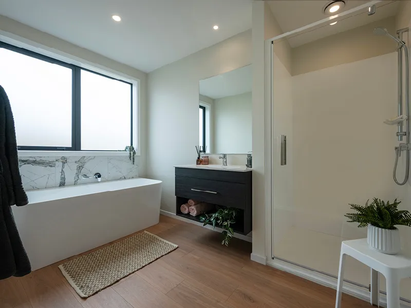 Modern Whitianga family bathroom with freestanding bathtub under frosted window, timber-look flooring, floating dark vanity and glass shower, styled with neutral tones and greenery accents.