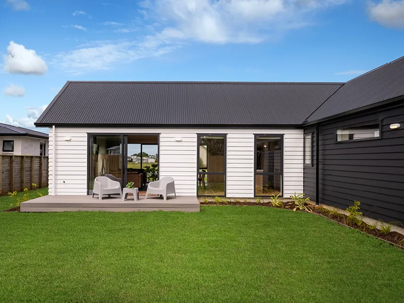 Modern Whitianga home backyard with white weatherboard exterior, sliding doors opening to raised deck with outdoor seating and landscaped lawn under blue sky.