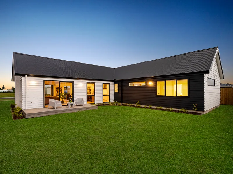 Modern Whitianga home exterior at dusk with L-shaped layout, white and black weatherboard cladding, raised deck with outdoor seating and expansive landscaped lawn.