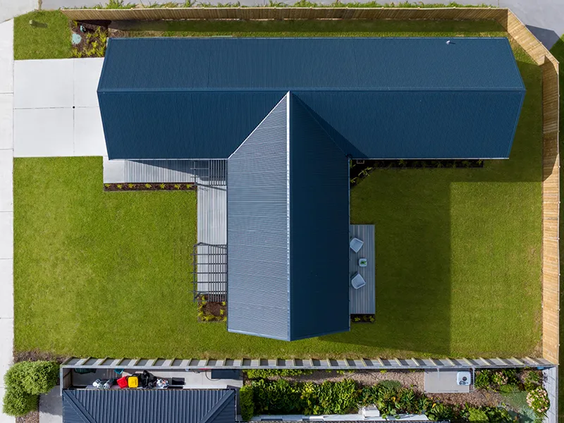 Aerial view of modern Whitianga home with dark gable roof, L-shaped layout, central deck and landscaped lawn enclosed by fencing and garden borders.