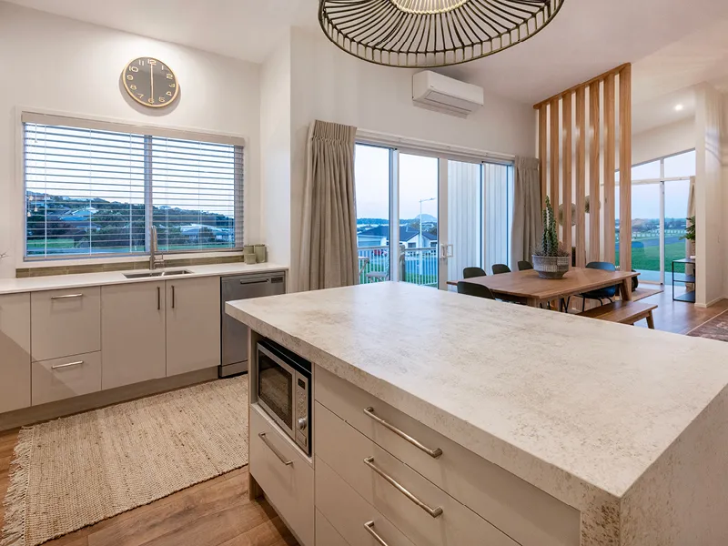Open-plan kitchen with stone island bench, wood-slat divider and dining area overlooking Ōmokoroa estuary in the Heart Foundation Home Lottery No. 165 house.