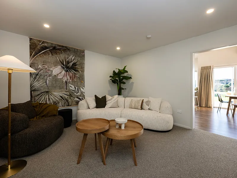 Living room with cream sofa, floral wall tapestry and round timber coffee tables opening onto the kitchen and dining area in the Heart Foundation Home Lottery No. 165 Ōmokoroa house.