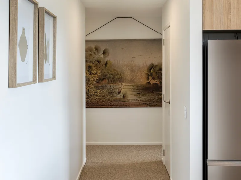 Hallway with wetland-themed wall tapestry, framed artwork and timber kitchen cabinetry in the Heart Foundation Home Lottery No. 165 Ōmokoroa house.