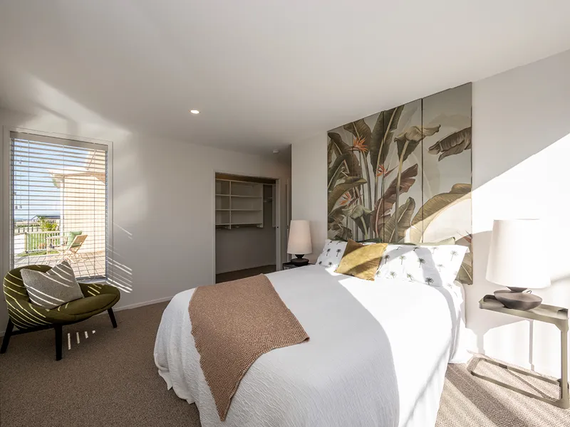 Sunlit bedroom with tropical wall art, olive armchair and walk-in wardrobe leading to a balcony with sea views in the Heart Foundation Home Lottery No. 165 Ōmokoroa house.