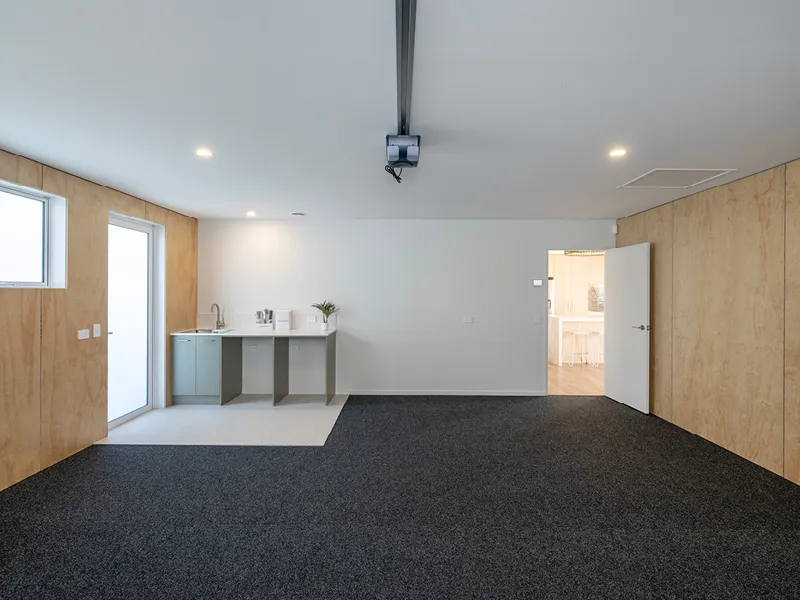 Multi-purpose flexi room with kitchenette, projector and dark carpet tiles opening to the main kitchen in the Heart Foundation Home Lottery No. 165 Ōmokoroa house.
