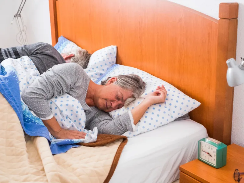 Older woman lying awake in bed at night looking uncomfortable while her partner sleeps beside her.