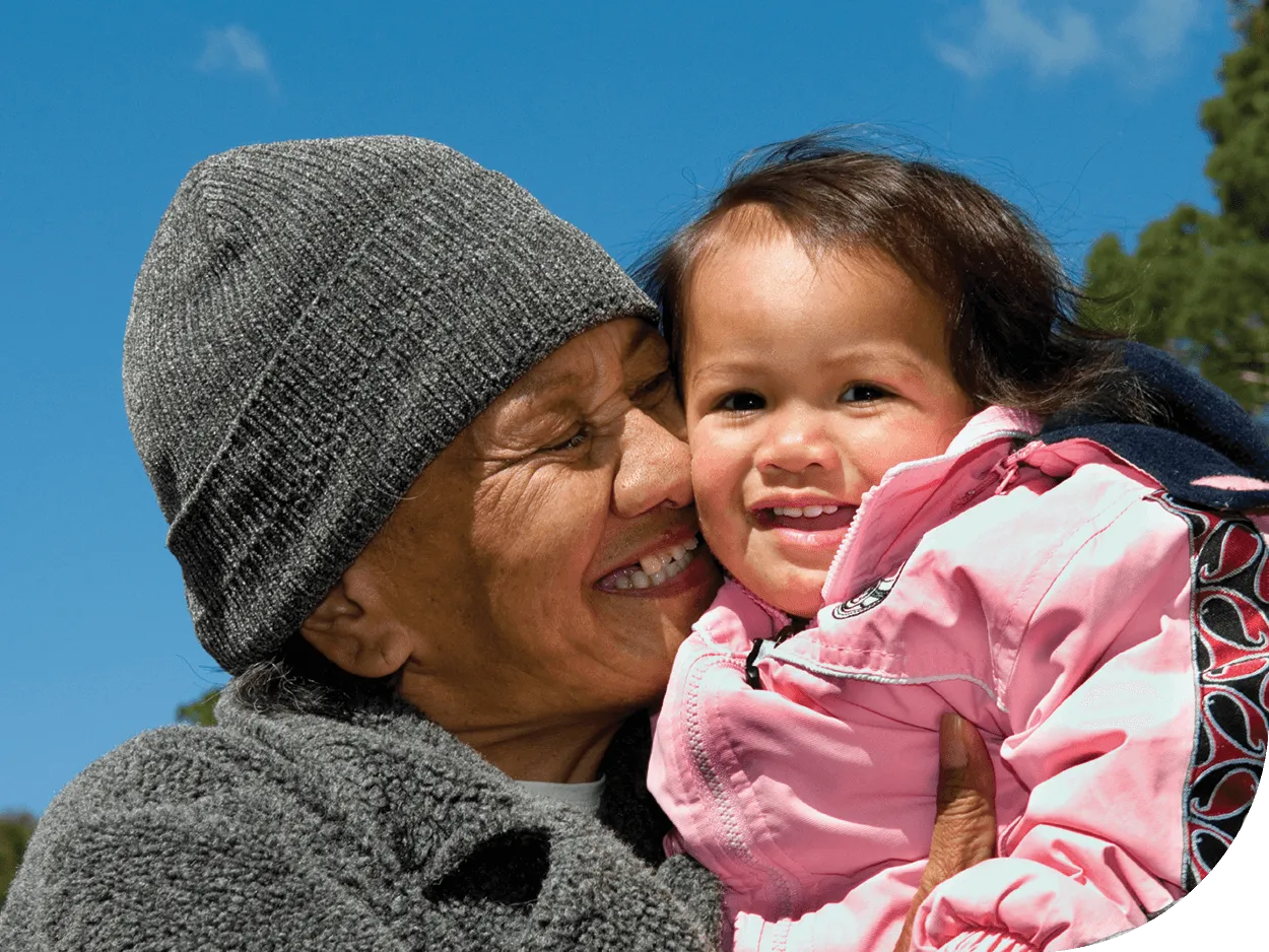 A senior Maori lady holds her baby granddaughter in an embrace. Both are smiling happily.