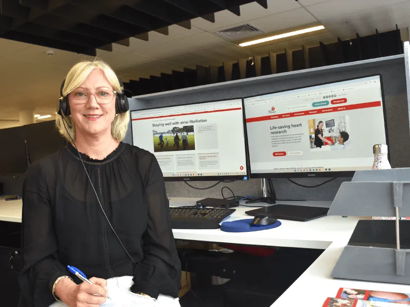 A woman wearing headphones sits at an office desk with dual computer monitors displaying heart health and atrial fibrillation information; she writes notes on paper in a modern workplace environment.