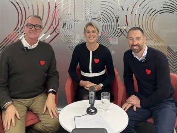 Three people wearing black sweaters with red heart logos sit together in a discussion setting with a microphone and water glasses on the table in front of them, against a modern patterned glass backdrop.