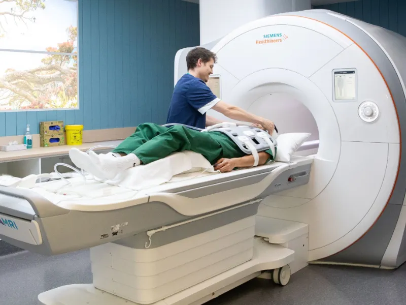 Radiographer positioning a patient on the table of a Siemens MRI scanner, preparing them for a cardiac MRI heart scan in a modern hospital imaging room.