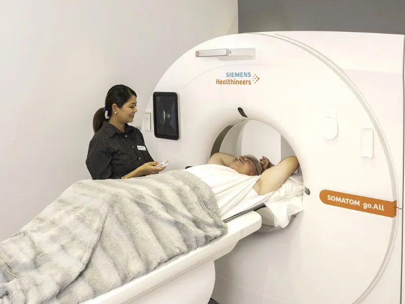 Radiographer standing beside a Siemens SOMATOM CT scanner while a patient lies on the table under a blanket, preparing for a coronary artery calcium scoring heart scan in a clinic.