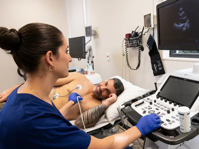 Image showing a clinician performing a transthoracic echocardiogram, using an ultrasound probe and ECG leads to capture live images of a patient’s heart on a monitor in a hospital or clinic setting.