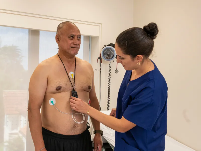 Clinician fitting a cardiac event monitor on a patient, attaching chest electrodes and demonstrating how to wear the small portable heart rhythm recorder during daily activities.