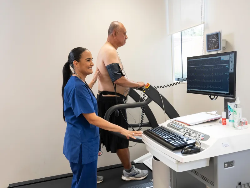Image showing a patient walking on a treadmill during an exercise tolerance test, while a clinician monitors the heart’s electrical activity and vital signs using ECG leads and equipment in a clinic setting.
