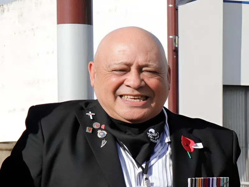 Older male veteran in blazer with service medals and remembrance poppy sitting outdoors at commemorative event.