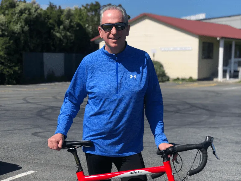 Chris Hooper in a blue long-sleeve athletic top and sunglasses standing outdoors with a red road bicycle on a sunny day. A building with a red roof and some greenery is visible in the background.