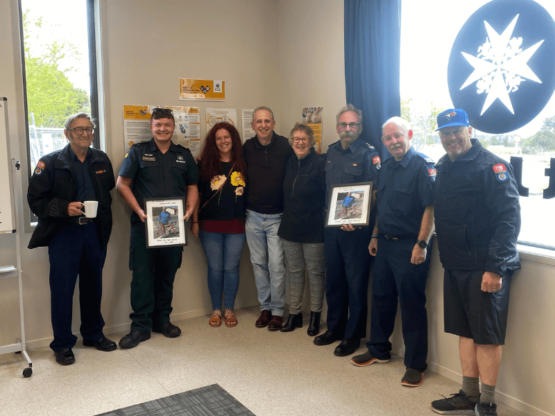 Group photo of eight people, including uniformed emergency service workers, standing together indoors. Two hold framed awards, while others stand casually in front of a window with an emergency services emblem and safety posters on the wall.