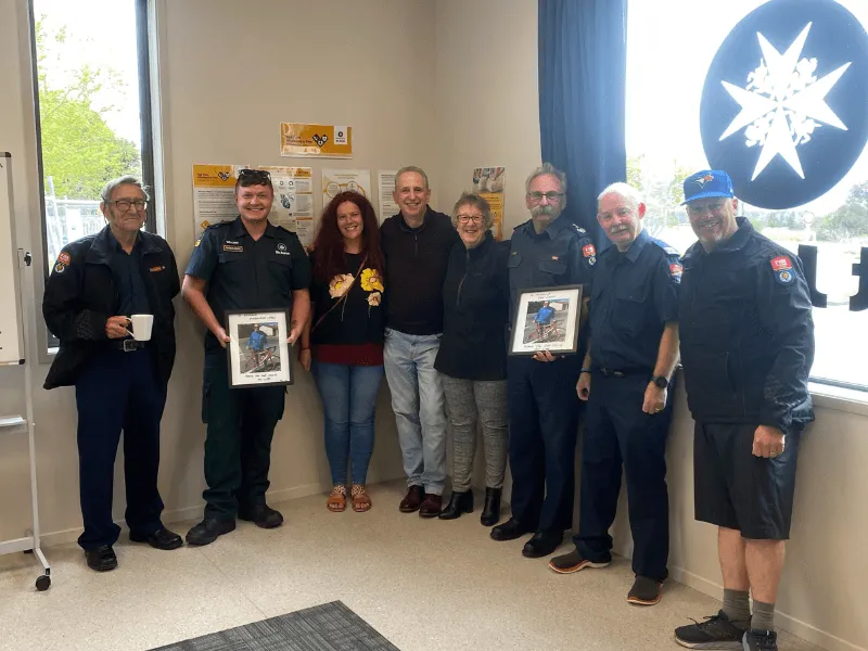 Group photo of eight people, including uniformed emergency service workers, standing together indoors. Two hold framed awards, while others stand casually in front of a window with an emergency services emblem and safety posters on the wall.