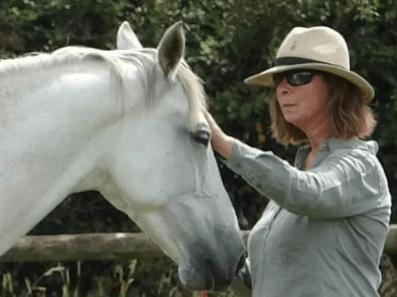 A woman wearing a wide-brimmed hat gently strokes a white horse outdoors near a wooden fence and greenery.