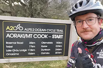 A head shot of Hamish Waugh wearing a cycle helmet and cycling clothes next to the Mount Cook alpine cycling trail sign. He is giving a thumbs up.