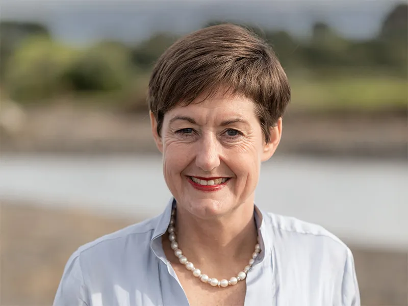 Portrait of Kara Northcott standing on a beach. She has short brown hair, is Caucasian in colouring and is smiling broadly at the camera. She is wearing a white button-down shirt and has a string of pearls at her neckline.