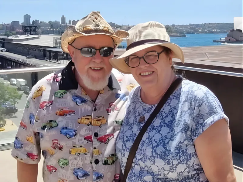 Couple on sunny waterfront balcony wearing summer hats, colorful retro car shirt and floral top with city skyline and blue harbor in background.
