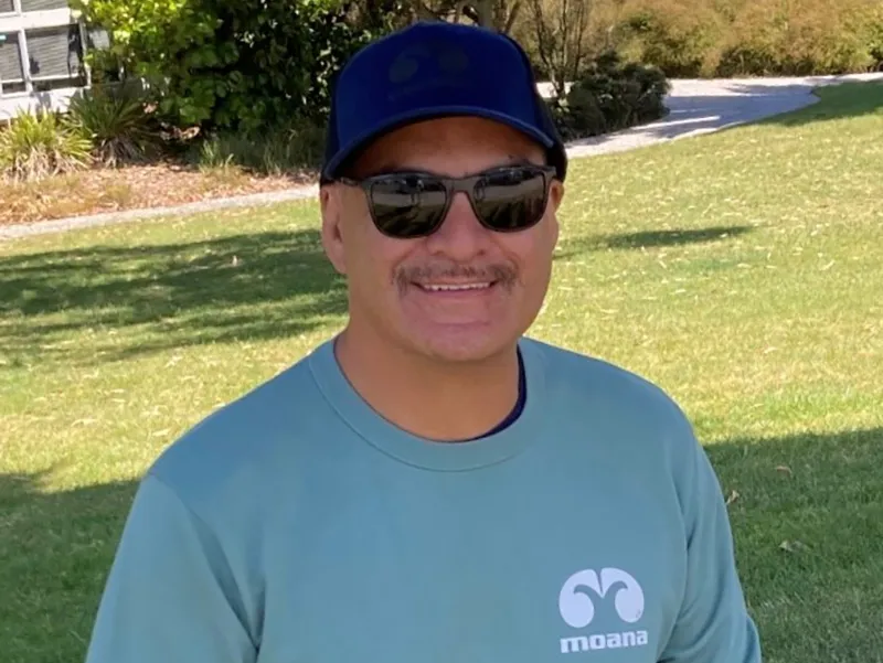 Photo of Kipa Rangiheuea sitting in the garden. He is wearing a baseball hat, sunglasses and a teal sweatshirt.