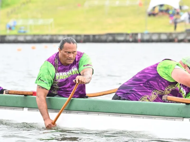 Waka ama paddler in purple and green team uniform powering canoe stroke on calm water during race.