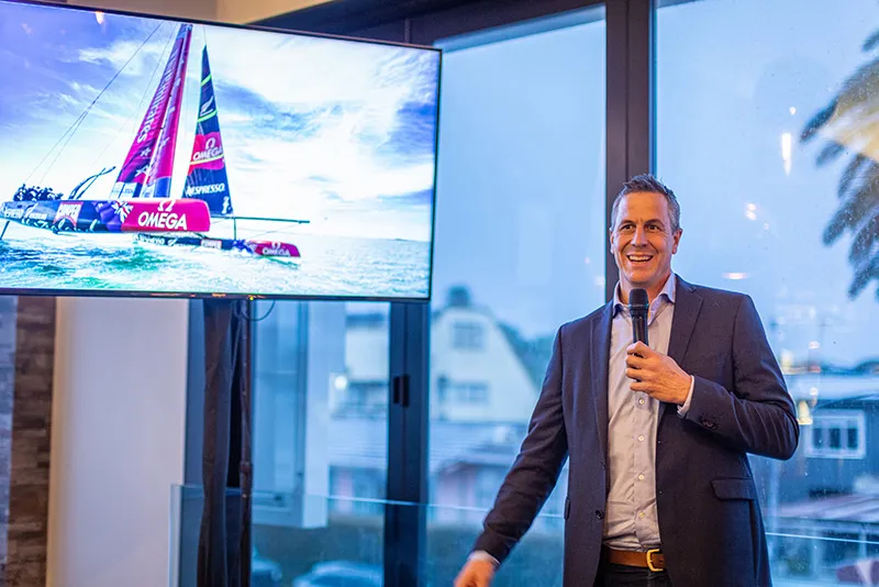 Image of Rob Waddell, Olympic Rower, speaking into a microphone whilst giving a presentation with a photo of an Americas Cup boat on a screen behind him.