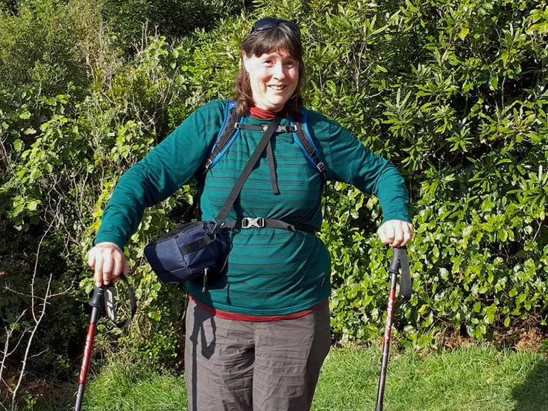 Sandra is a middle-aged women with brown shoulder-length hair. She is standing outside surrounded by bushes and she has two walking sticks - one in each hand. She is wearing hiking clothes.