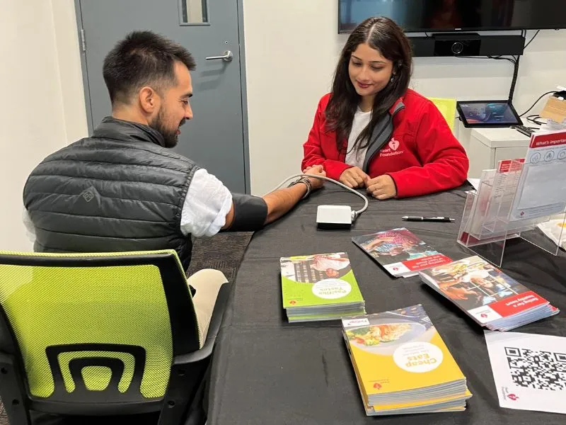 “Heart Foundation Heart Health Promoter taking employee blood pressure at New Zealand workplace wellness session with heart health resources on table”.
