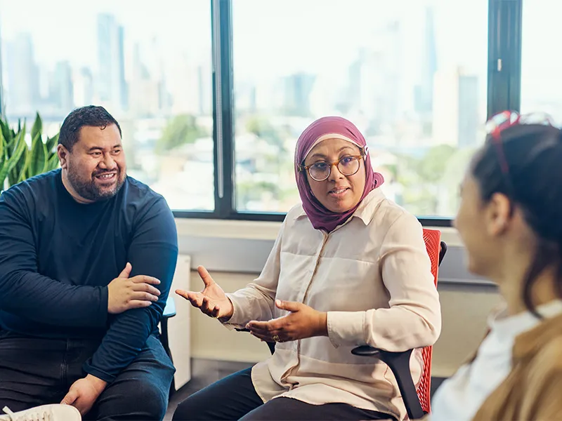 Three people sit together in a bright office space having a discussion, with one person gesturing while talking and a large city window view in the background.