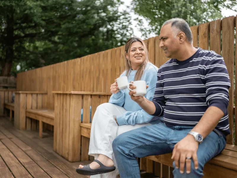 Two people are sitting outdoors on wooden benches, enjoying hot drinks together in a relaxed setting surrounded by trees and a tall wooden fence.