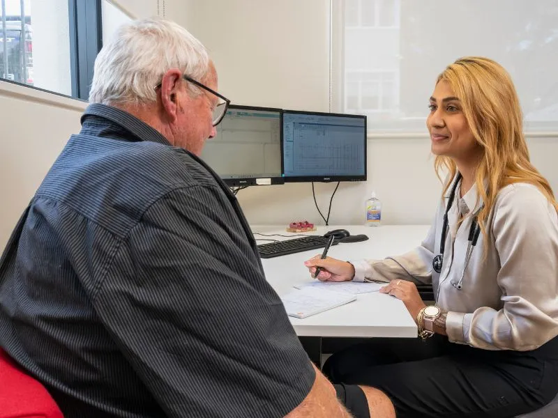 Doctor consulting elderly male patient in a modern medical office, with computer monitors in the background and patient records visible on the desk.