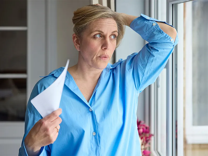 Middle-aged menopausal woman in a blue shirt at home having a hot flush. She is cooling herself with a piece of paper, fanning in front of her face. With the other hand she holds her hair away from her neck.
